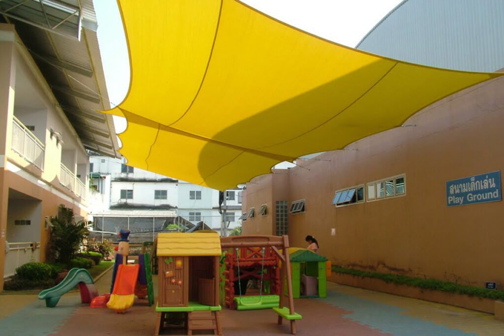 Cánh buồm vải vàng căng giữa dãy lớp học và tường, bên dưới là nhà chơi gỗ, cầu trượt và biển Play Ground