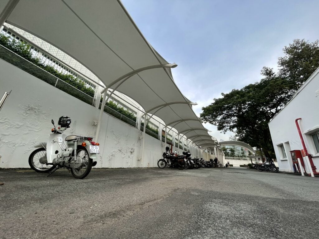 Bãi xe máy dưới dãy mái màng căng trắng nhìn từ mặt đất, xe máy đỗ sát tường, cây lớn và nhà trắng bên phải