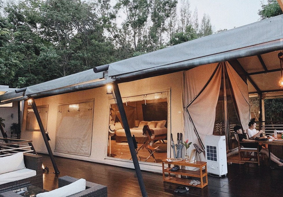 Hiên villa glamping dưới mái màng căng xám, rèm vải mở ra phòng ngủ, khách ngồi bàn ăn ngoài hiên