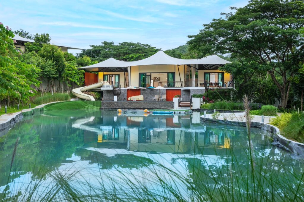 Villa glamping mái màng căng ba đỉnh trắng soi bóng xuống hồ bơi tự nhiên, cây xanh và trời xanh