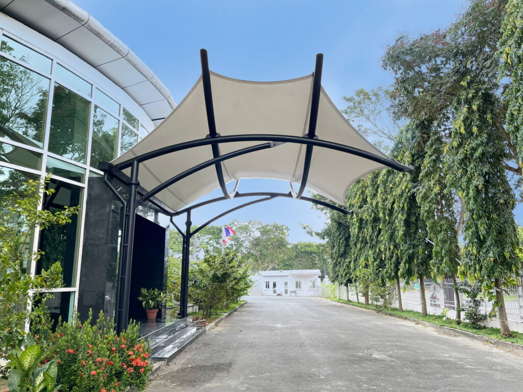 Mái che lối vào nhà xưởng hình cánh diều: màng trắng trên khung thép đen cong trước sảnh kính, đường nội bộ hai hàng cây