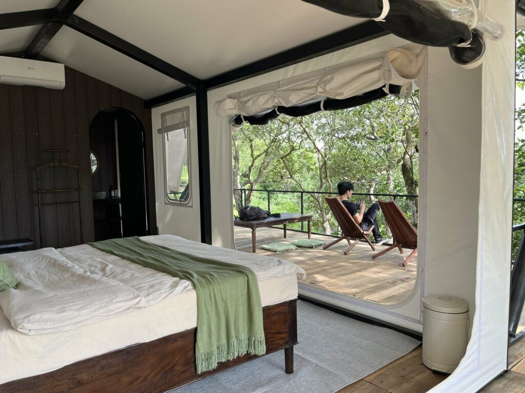 Phòng ngủ trong lều glamping, cửa bạt cuộn lên mở ra hiên gỗ có ghế nằm giữa rừng