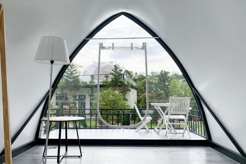 Cửa vòm lớn của lều glamping mở ra hiên có bàn ghế trắng, lan can thép và vườn cây, đèn cây chao trắng trong phòng