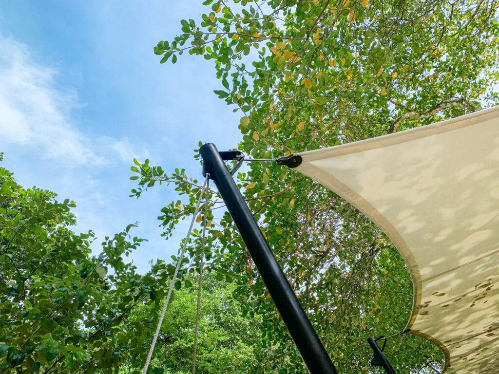 Cột thép đen nghiêng có dây néo, góc màng căng trắng nối vào đầu cột bằng phụ kiện inox, tán cây phía trên