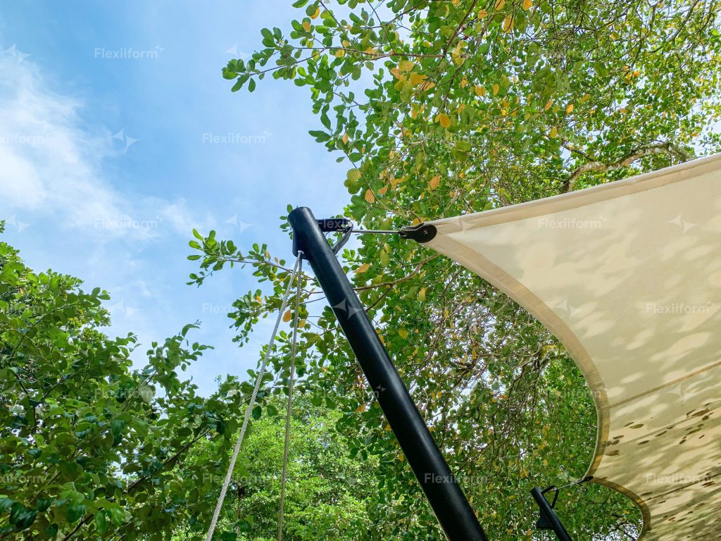 Cột thép đen nghiêng có dây néo, góc màng căng trắng nối vào đầu cột bằng phụ kiện inox, tán cây phía trên