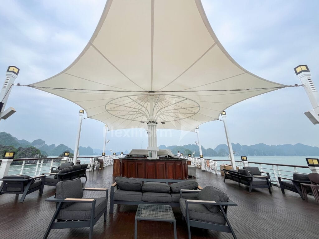 Mái che du thuyền vịnh Lan Hạ dạng chóp màng trắng trên quầy bar gỗ boong thượng, sofa xám, núi đá vôi phía xa
