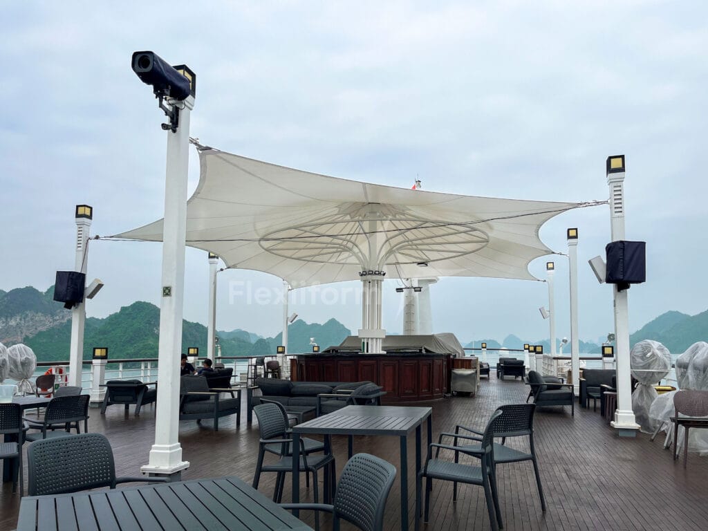 Boong thượng du thuyền với bàn ghế, quầy bar dưới chóp màng căng trắng, cột đèn và loa quanh lan can